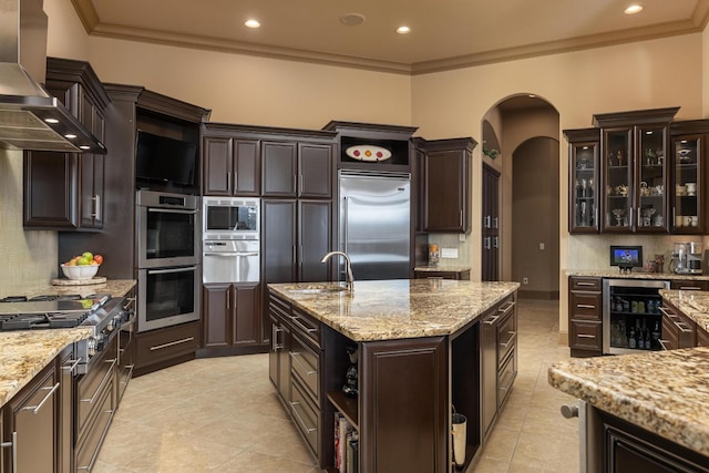 kitchen with sink, built in appliances, dark brown cabinetry, beverage cooler, and wall chimney exhaust hood