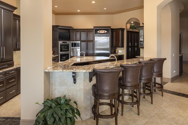 kitchen featuring a kitchen breakfast bar, built in appliances, light stone counters, dark brown cabinetry, and ornamental molding