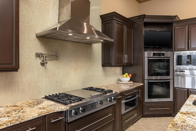 kitchen featuring light tile patterned flooring, light stone counters, wall chimney range hood, stainless steel appliances, and backsplash