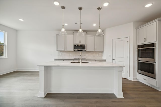 kitchen featuring stainless steel appliances, decorative light fixtures, light stone countertops, and a center island with sink