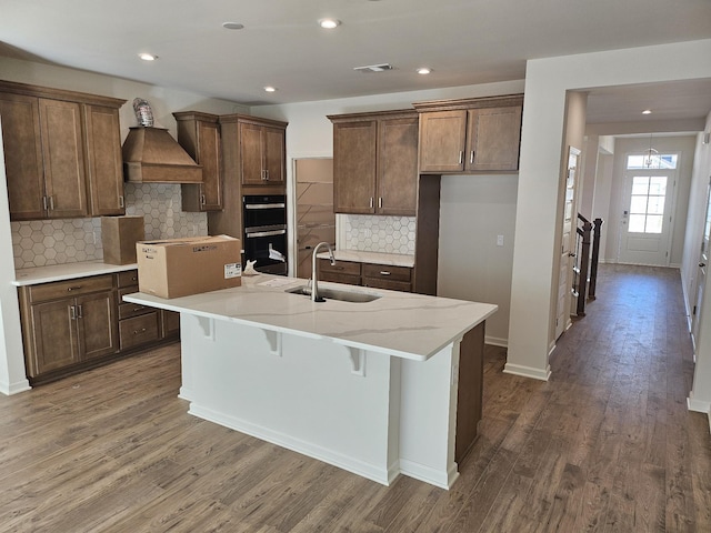 kitchen featuring premium range hood, sink, dark hardwood / wood-style floors, an island with sink, and black double oven