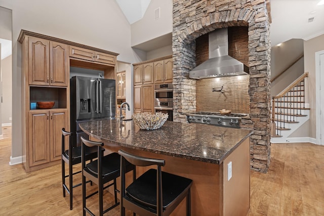 kitchen with wall chimney range hood, a breakfast bar area, a kitchen island with sink, stainless steel appliances, and light hardwood / wood-style floors