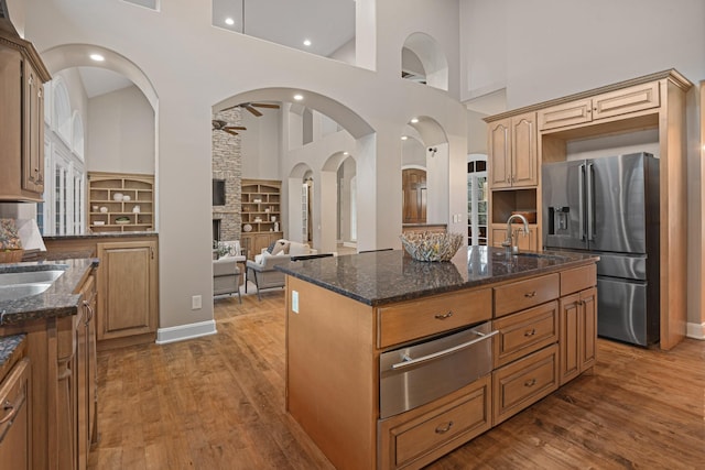 kitchen featuring wood-type flooring, an island with sink, dark stone counters, ceiling fan, and stainless steel refrigerator with ice dispenser