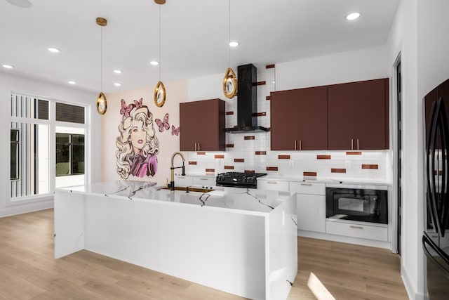 kitchen featuring decorative light fixtures, wall chimney range hood, light hardwood / wood-style floors, black appliances, and a center island with sink