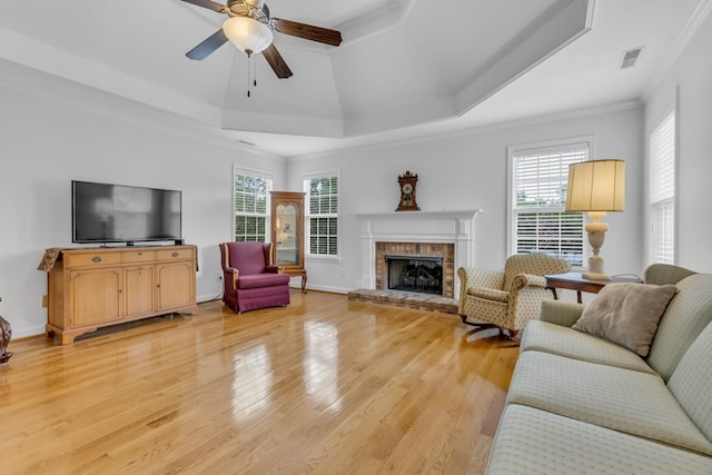 living room featuring light wood-type flooring, ornamental molding, a tray ceiling, ceiling fan, and a fireplace