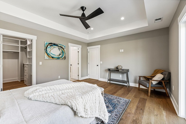 bedroom with a raised ceiling, wood-type flooring, a walk in closet, and ceiling fan