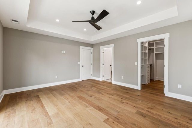 unfurnished bedroom with a walk in closet, a tray ceiling, light hardwood / wood-style flooring, and ceiling fan