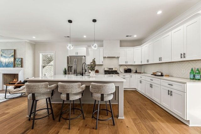 kitchen with premium range hood, decorative light fixtures, a center island with sink, stainless steel appliances, and white cabinets