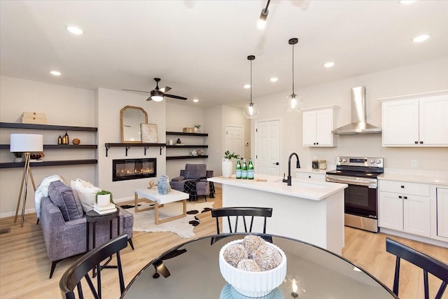 kitchen with wall chimney exhaust hood, hanging light fixtures, electric range, a kitchen island with sink, and white cabinets