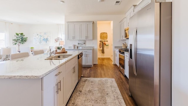 kitchen with appliances with stainless steel finishes, wood-type flooring, sink, light stone counters, and a center island with sink