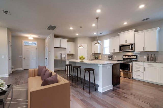 kitchen with pendant lighting, white cabinetry, light stone counters, stainless steel appliances, and a center island with sink