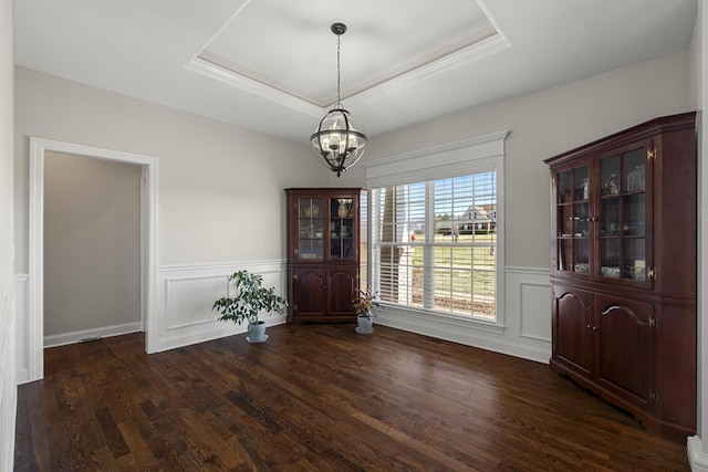 unfurnished dining area with a tray ceiling, a wainscoted wall, a decorative wall, an inviting chandelier, and dark wood-type flooring