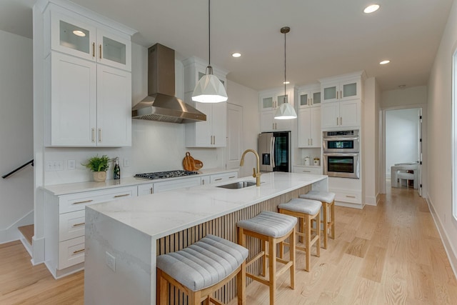 kitchen with an island with sink, sink, white cabinets, stainless steel appliances, and wall chimney range hood