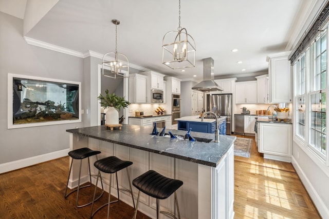 kitchen featuring appliances with stainless steel finishes, white cabinetry, island range hood, an island with sink, and decorative light fixtures