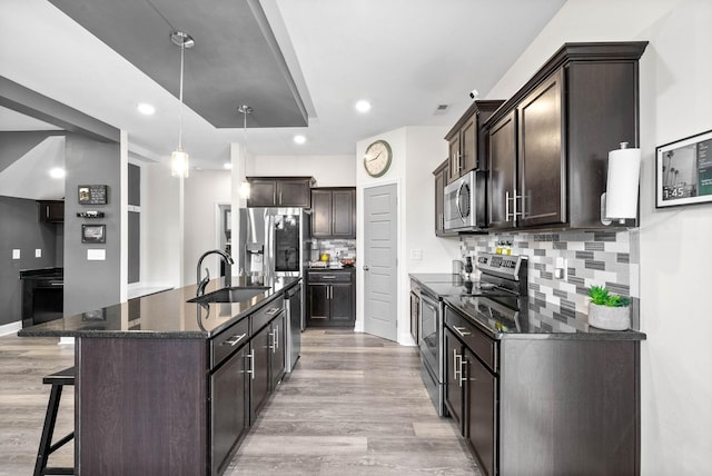 kitchen with appliances with stainless steel finishes, pendant lighting, dark stone countertops, a kitchen island with sink, and light hardwood / wood-style flooring