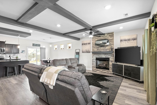 living room with beamed ceiling, a stone fireplace, ceiling fan, and light hardwood / wood-style flooring