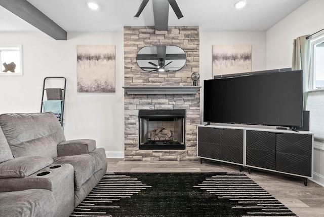 living room with beam ceiling, light hardwood / wood-style flooring, a stone fireplace, and ceiling fan