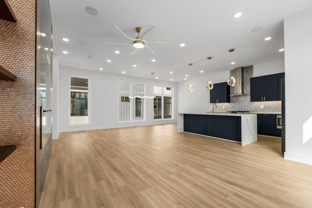 kitchen featuring a kitchen island with sink, wall chimney range hood, hanging light fixtures, and light wood-type flooring