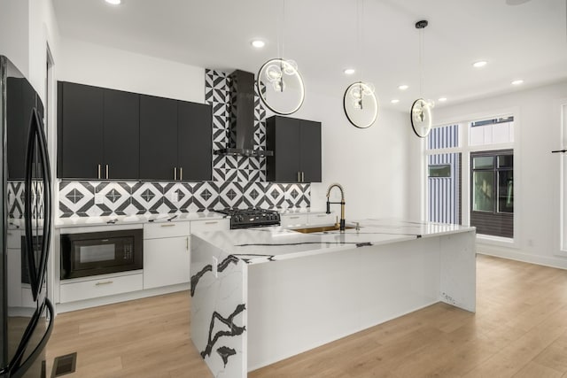 kitchen with pendant lighting, an island with sink, light hardwood / wood-style floors, black appliances, and wall chimney range hood