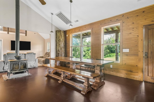 dining space featuring wooden walls, high vaulted ceiling, ceiling fan, and a wood stove