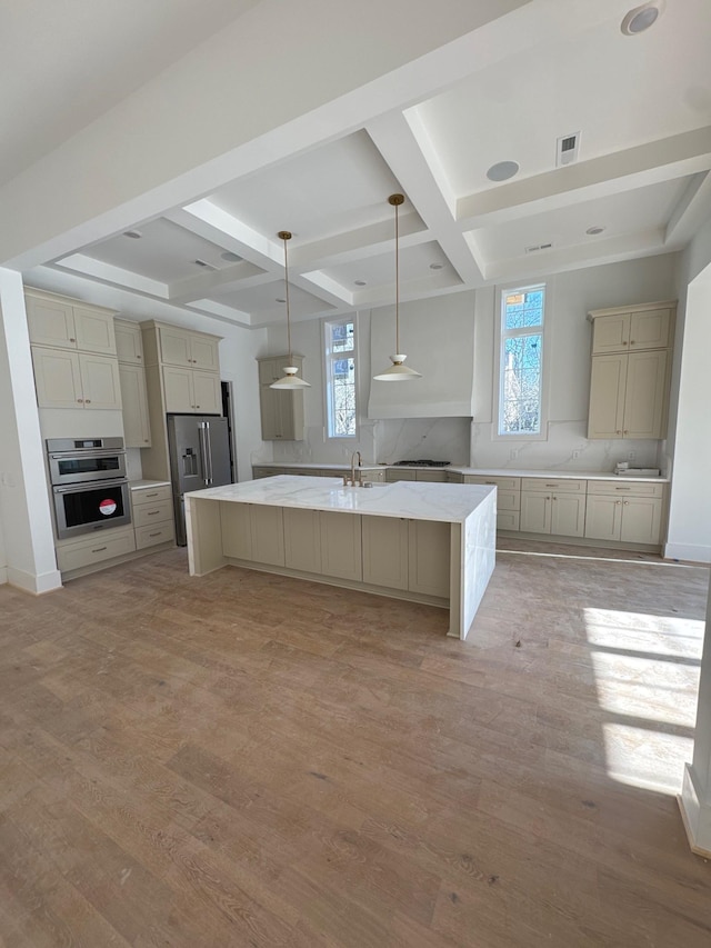 kitchen with an island with sink, pendant lighting, stainless steel appliances, light stone countertops, and light hardwood / wood-style floors