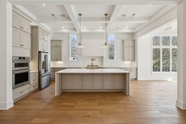 kitchen featuring appliances with stainless steel finishes, a kitchen island with sink, hanging light fixtures, coffered ceiling, and light hardwood / wood-style floors