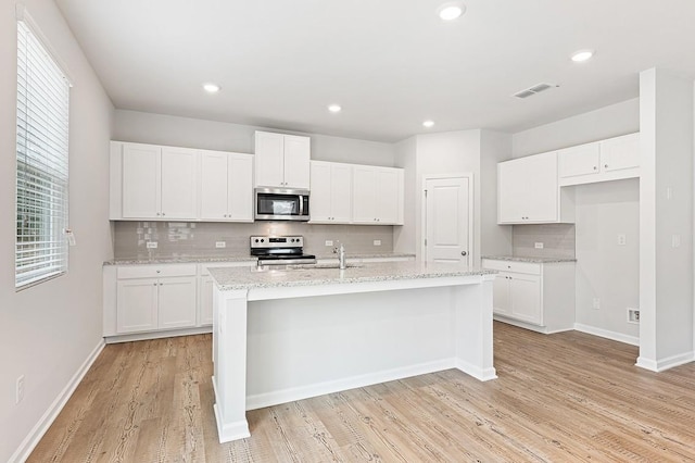 kitchen with appliances with stainless steel finishes, white cabinetry, light stone countertops, a center island with sink, and light wood-type flooring