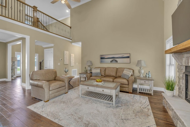 living room with ceiling fan, a high ceiling, ornamental molding, dark hardwood / wood-style flooring, and a stone fireplace