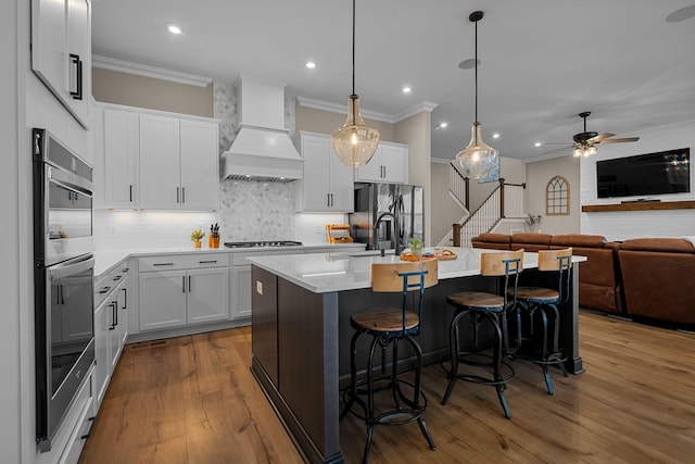 kitchen with a breakfast bar, white cabinets, stainless steel appliances, custom range hood, and a center island with sink