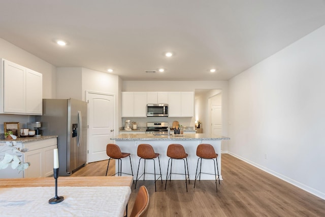 kitchen featuring appliances with stainless steel finishes, hardwood / wood-style floors, white cabinetry, light stone counters, and a center island with sink