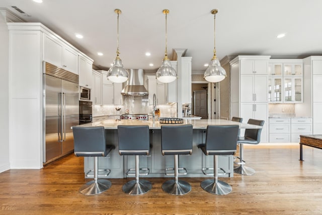 kitchen with built in appliances, wall chimney range hood, decorative light fixtures, and white cabinetry