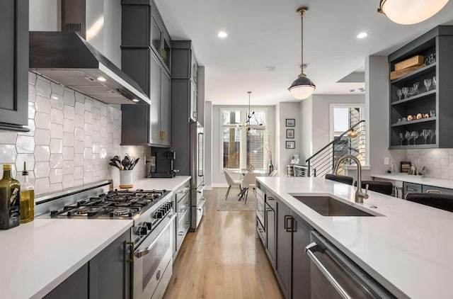 kitchen with pendant lighting, stainless steel appliances, a sink, wall chimney range hood, and light wood-type flooring