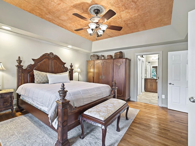 bedroom featuring connected bathroom, light hardwood / wood-style flooring, ceiling fan, and a tray ceiling