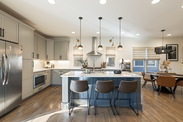 kitchen with gray cabinetry, a center island with sink, appliances with stainless steel finishes, pendant lighting, and wall chimney range hood