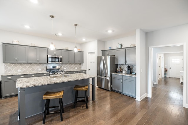 kitchen with dark hardwood / wood-style floors, decorative light fixtures, stainless steel appliances, light stone countertops, and a center island with sink