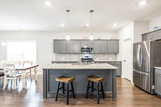 kitchen featuring pendant lighting, gray cabinets, appliances with stainless steel finishes, light stone counters, and an island with sink