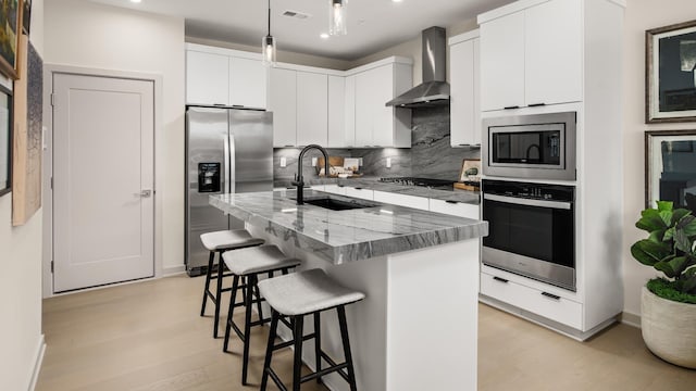 kitchen with sink, appliances with stainless steel finishes, an island with sink, wall chimney range hood, and white cabinets