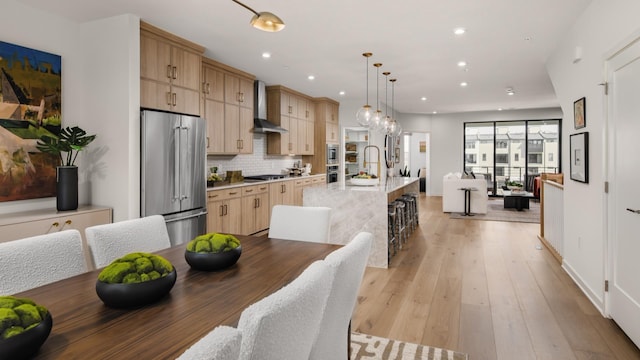 kitchen with wall chimney exhaust hood, light brown cabinetry, hanging light fixtures, a center island with sink, and stainless steel appliances