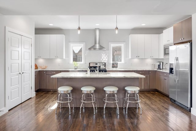 kitchen with wall chimney range hood, a breakfast bar, appliances with stainless steel finishes, an island with sink, and decorative light fixtures