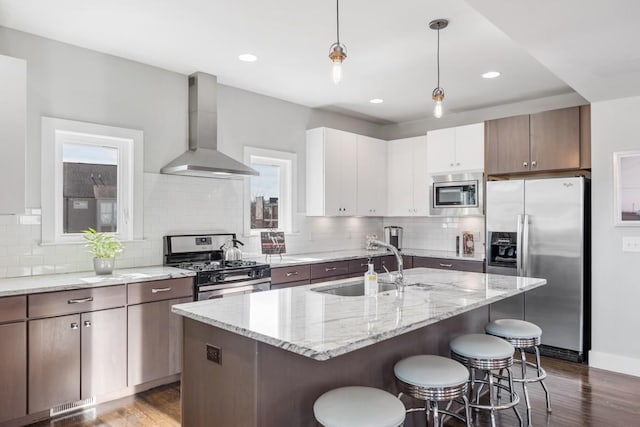 kitchen with wall chimney range hood, appliances with stainless steel finishes, white cabinetry, a center island with sink, and decorative light fixtures