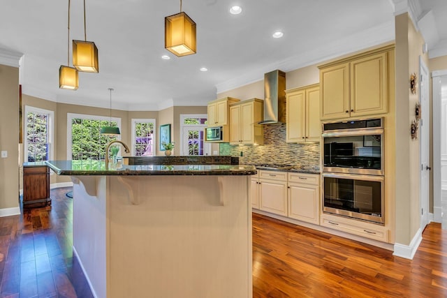 kitchen featuring appliances with stainless steel finishes, a kitchen bar, hanging light fixtures, a center island with sink, and wall chimney exhaust hood