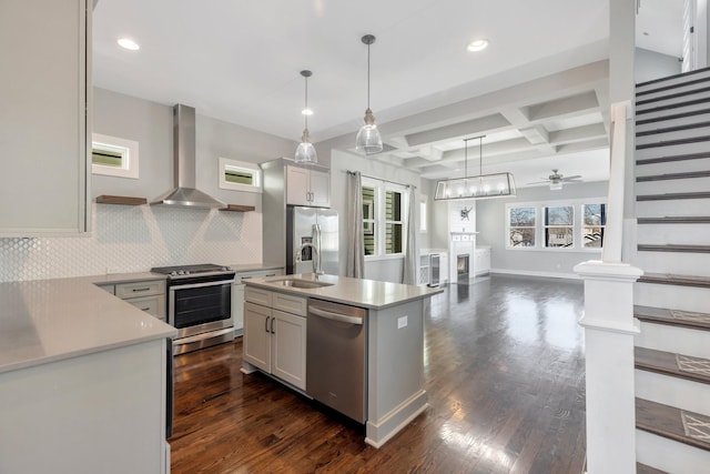 kitchen featuring appliances with stainless steel finishes, pendant lighting, an island with sink, coffered ceiling, and wall chimney range hood