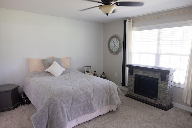 bedroom with a stone fireplace, light colored carpet, and ceiling fan