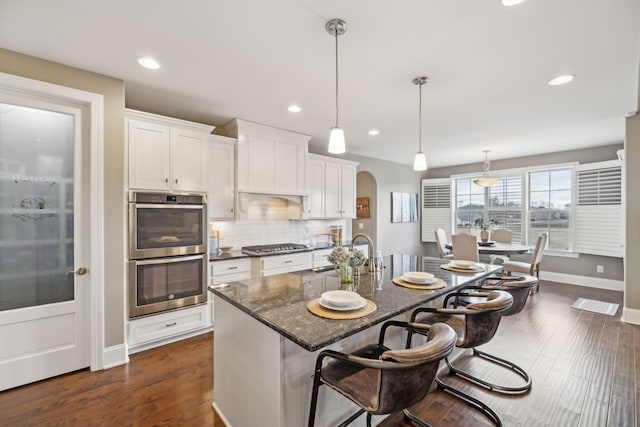 kitchen featuring white cabinets, dark stone counters, hanging light fixtures, a kitchen island with sink, and stainless steel appliances