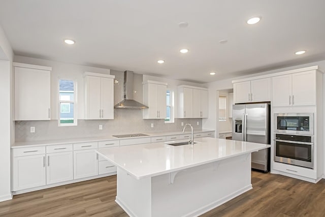 kitchen featuring wall chimney exhaust hood, sink, white cabinetry, appliances with stainless steel finishes, and an island with sink