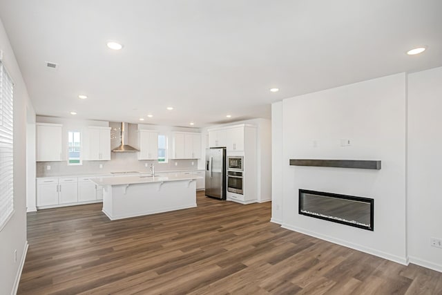 kitchen featuring wall chimney range hood, stainless steel appliances, an island with sink, white cabinets, and a kitchen bar