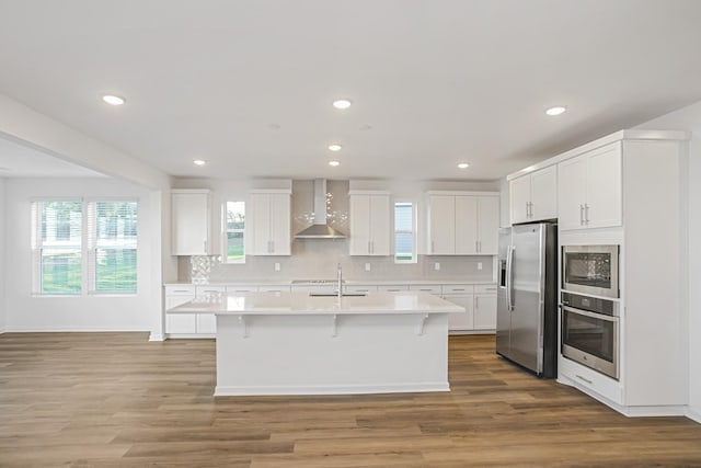 kitchen featuring appliances with stainless steel finishes, an island with sink, hardwood / wood-style flooring, wall chimney range hood, and white cabinets