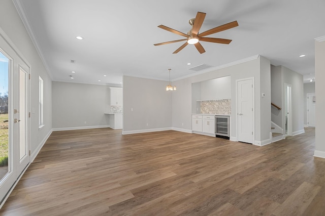 unfurnished living room featuring wine cooler, crown molding, ceiling fan with notable chandelier, and light wood-type flooring