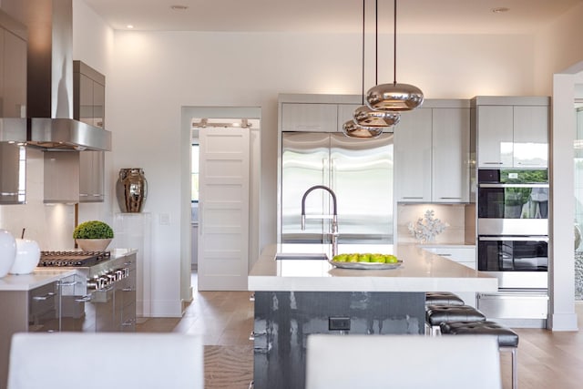 kitchen featuring wall chimney exhaust hood, appliances with stainless steel finishes, an island with sink, pendant lighting, and decorative backsplash