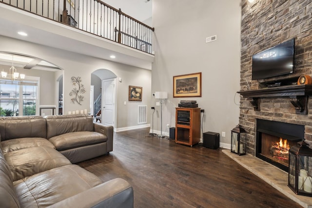 living room with dark wood-type flooring, a towering ceiling, a stone fireplace, and an inviting chandelier